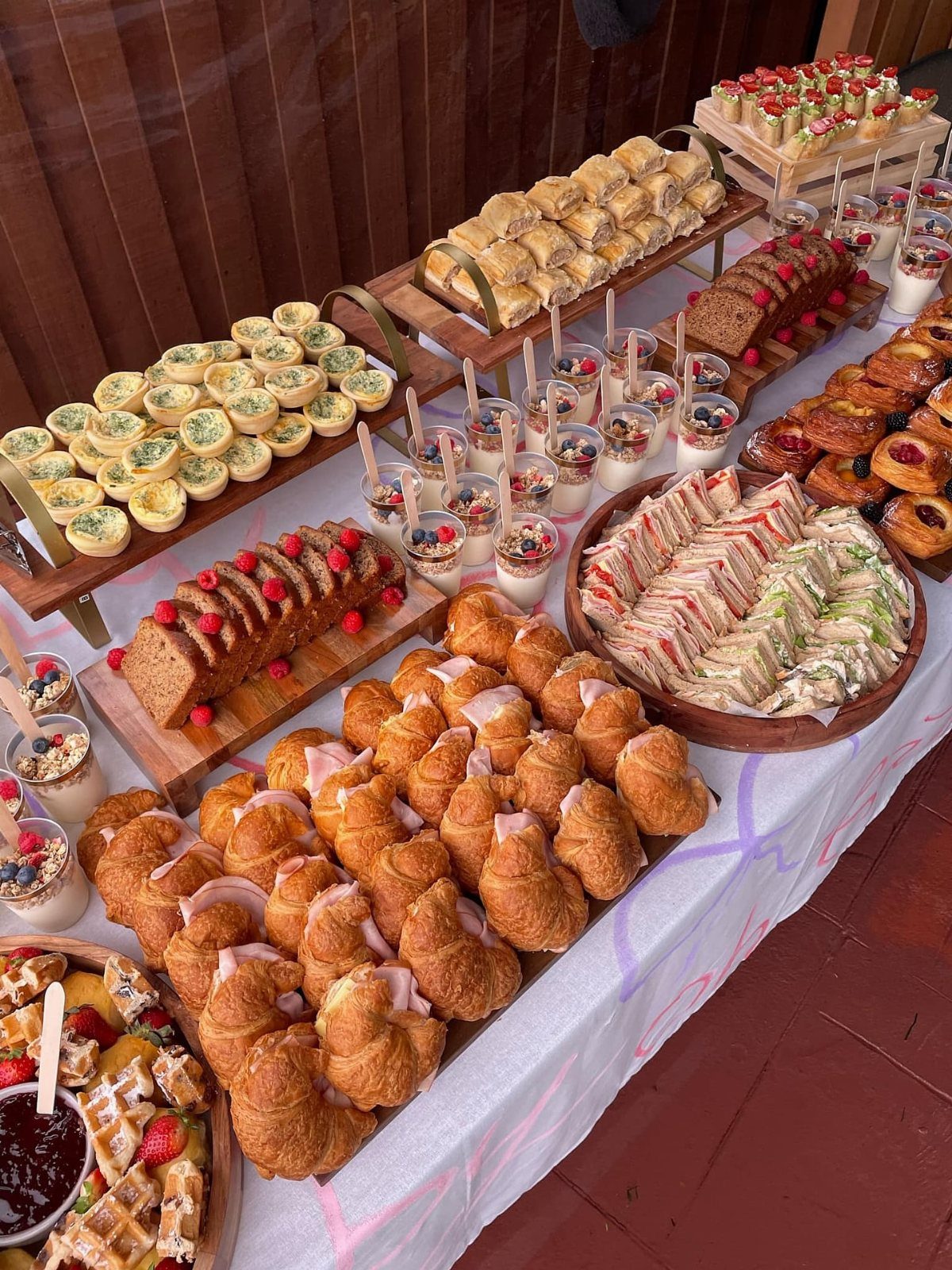 Breakfast buffet table with croissants, mini quiches, sandwiches, and fruit on white cloth.