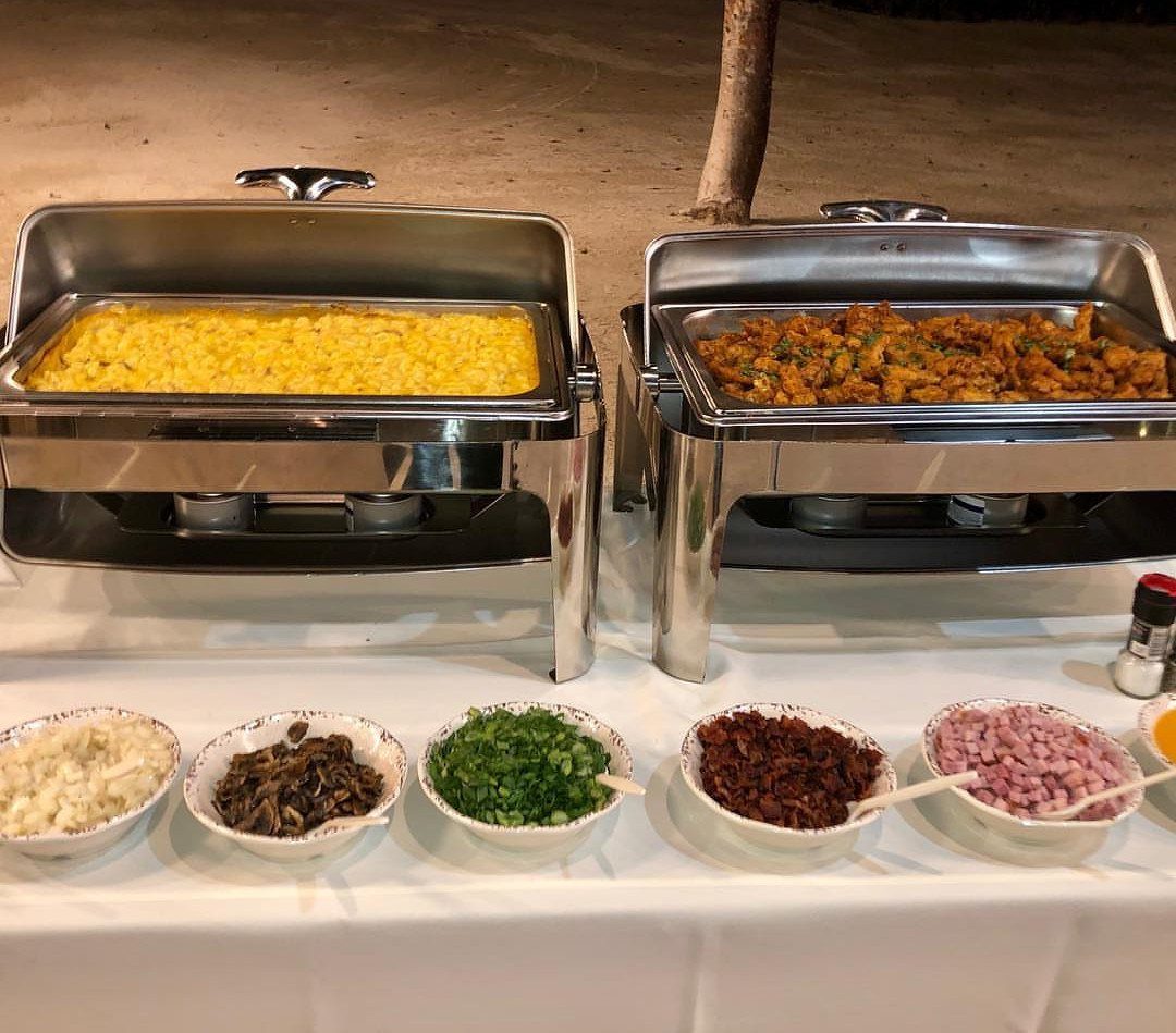 Macaroni and cheese in a chafing dish with various topping bowls on a table.