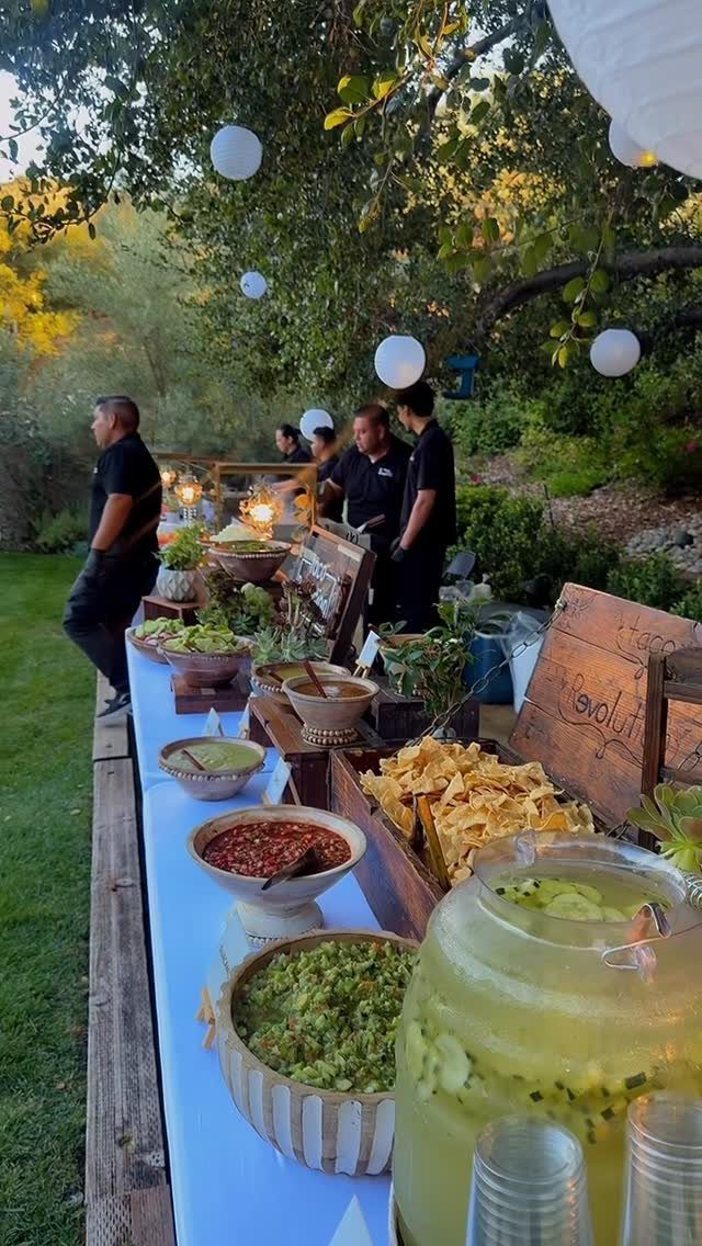 Outdoor wedding food station with dips, chips, and drinks under white globe lanterns.