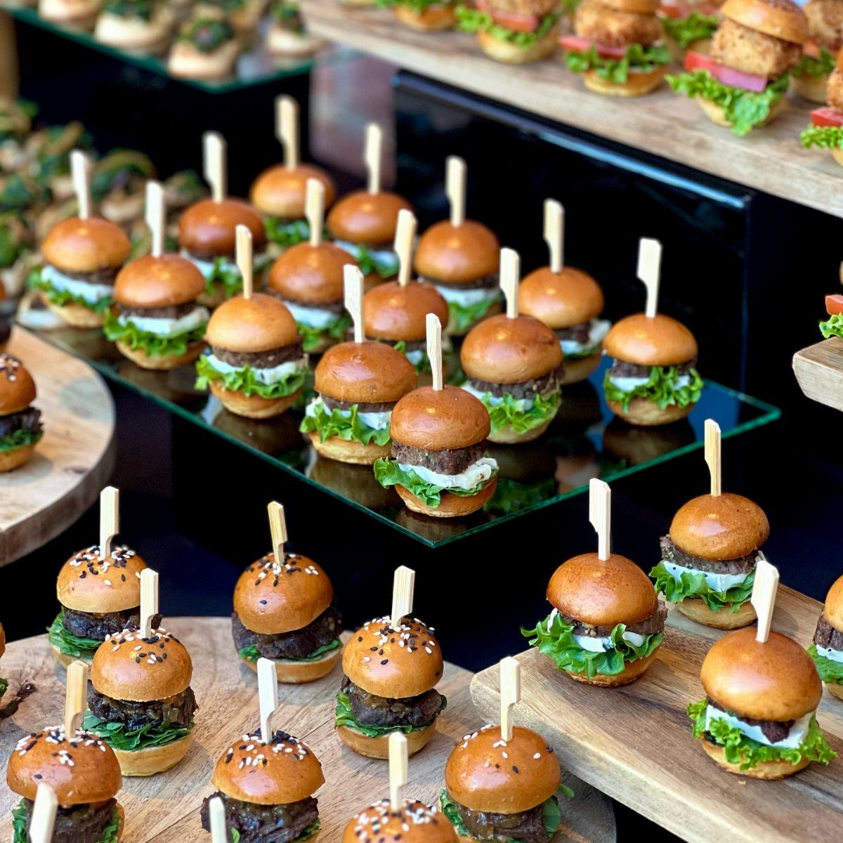 Miniature burgers with lettuce and sauce arranged on wooden and glass platters.