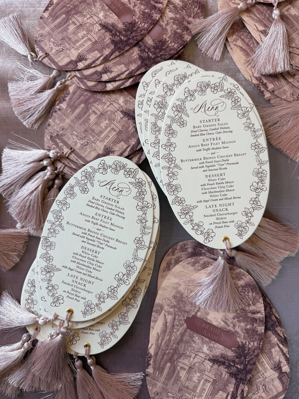 Oval white wedding menus with floral borders and mauve tassels on grey fabric