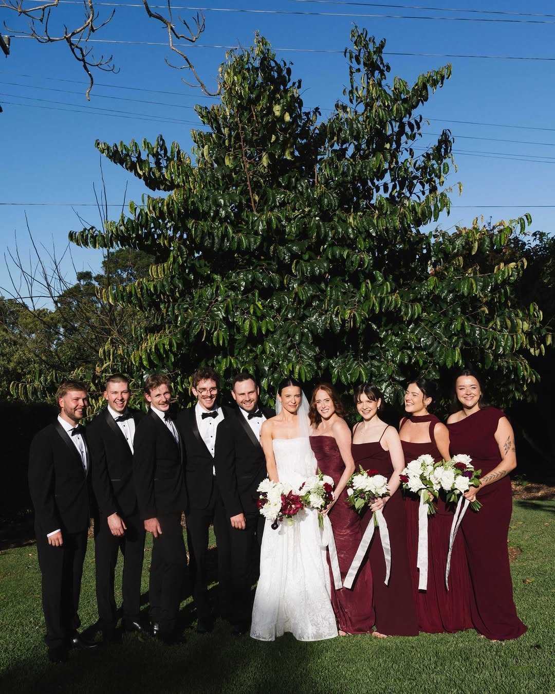 Wedding party posing outdoors with a bride in white and bridesmaids in burgundy gowns.