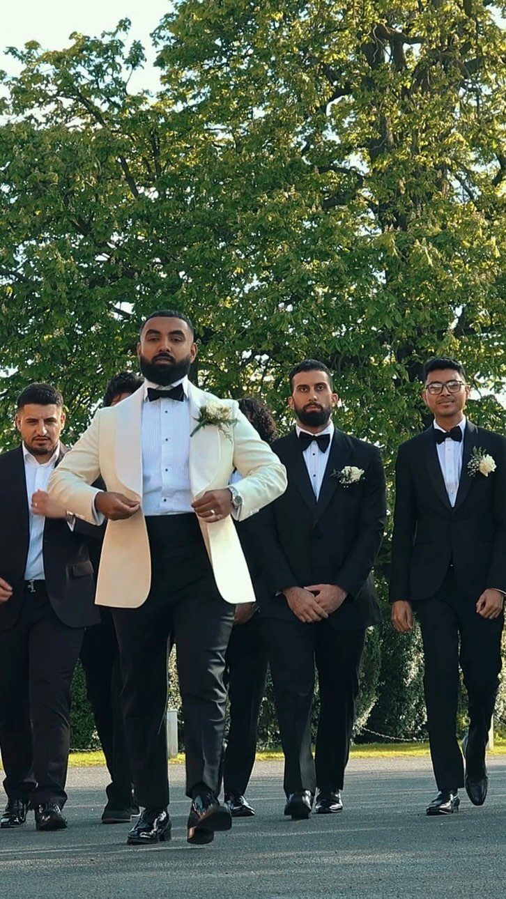 Groom in white tuxedo jacket walking with groomsmen in black tuxedos outdoors.
