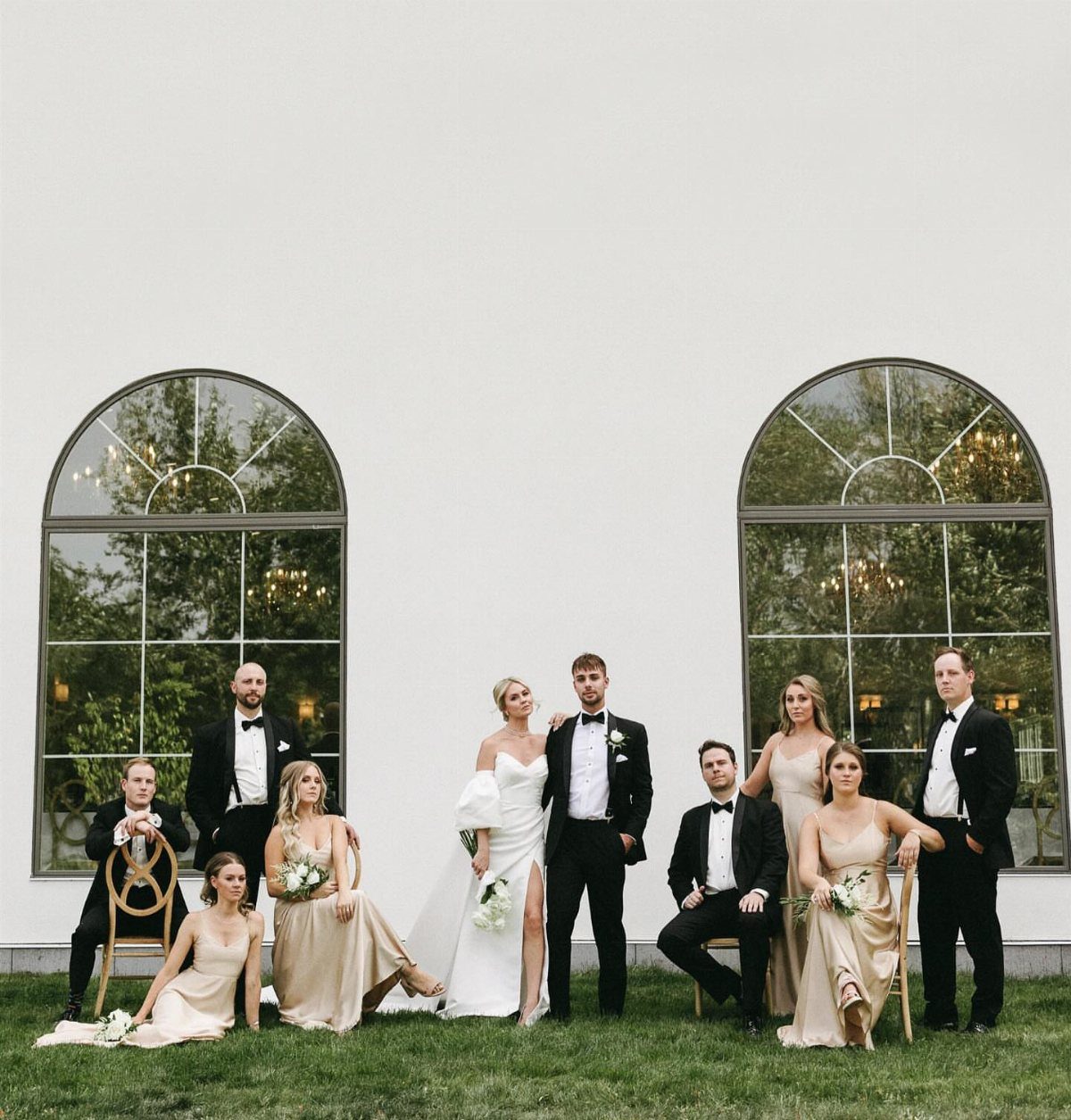 Wedding party posing formally in front of a white wall with arched windows.
