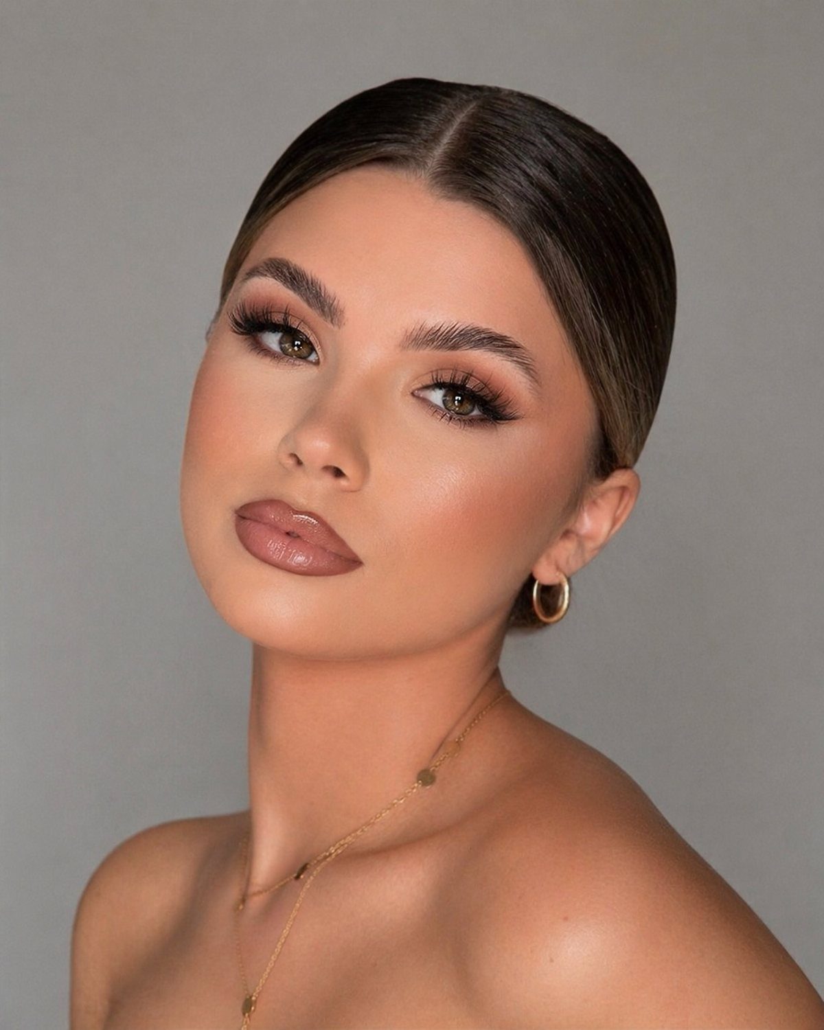 Woman with beige-pink glossy lips and warm glam makeup posing against grey background