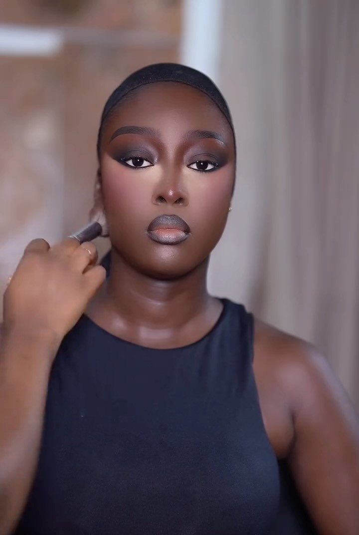 Woman with dark eyeshadow and neutral lips getting makeup applied to her cheek