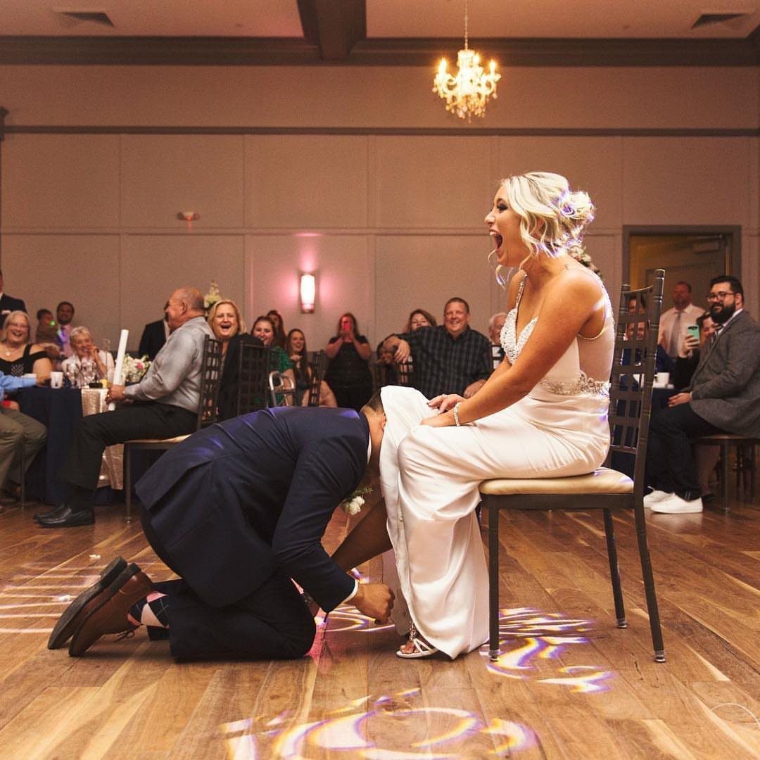 Bride laughing while groom kneels to remove her garter at a wedding reception.