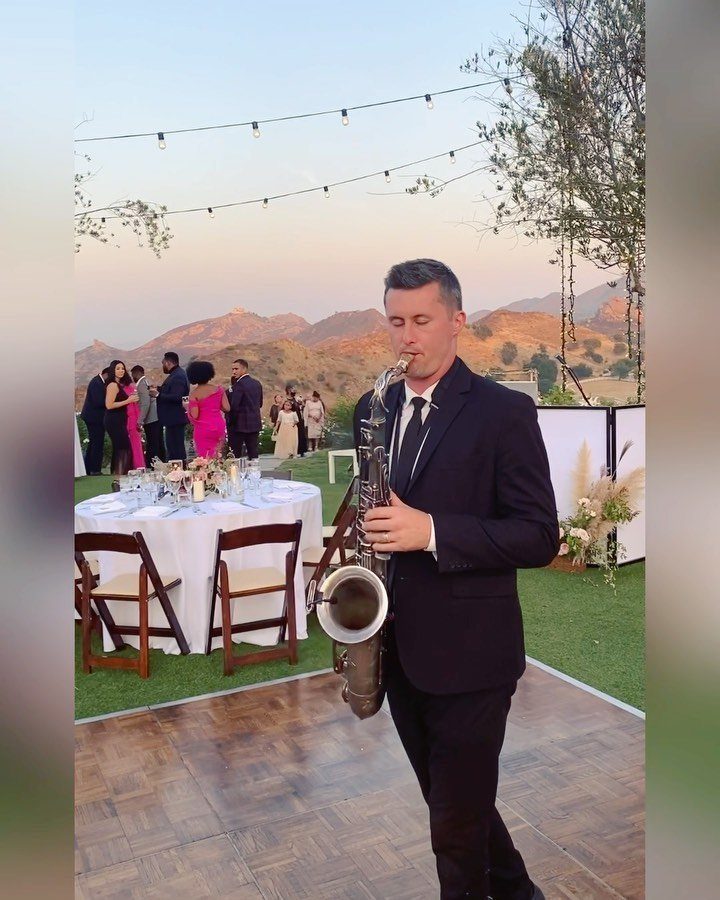 Saxophonist in a black suit playing music at an outdoor wedding reception.