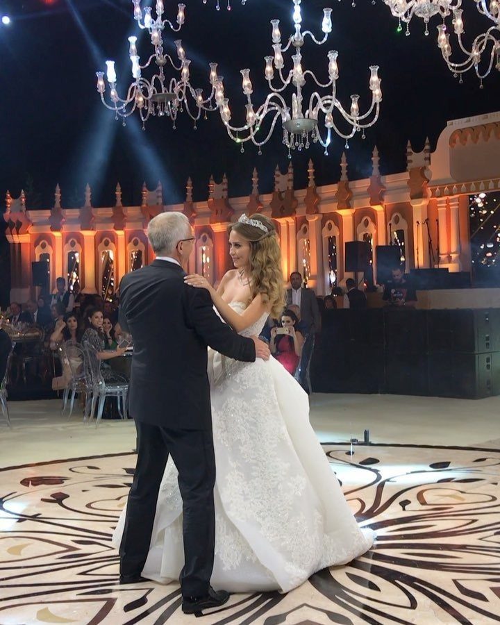Bride in white gown dancing with her father under crystal chandeliers at night.