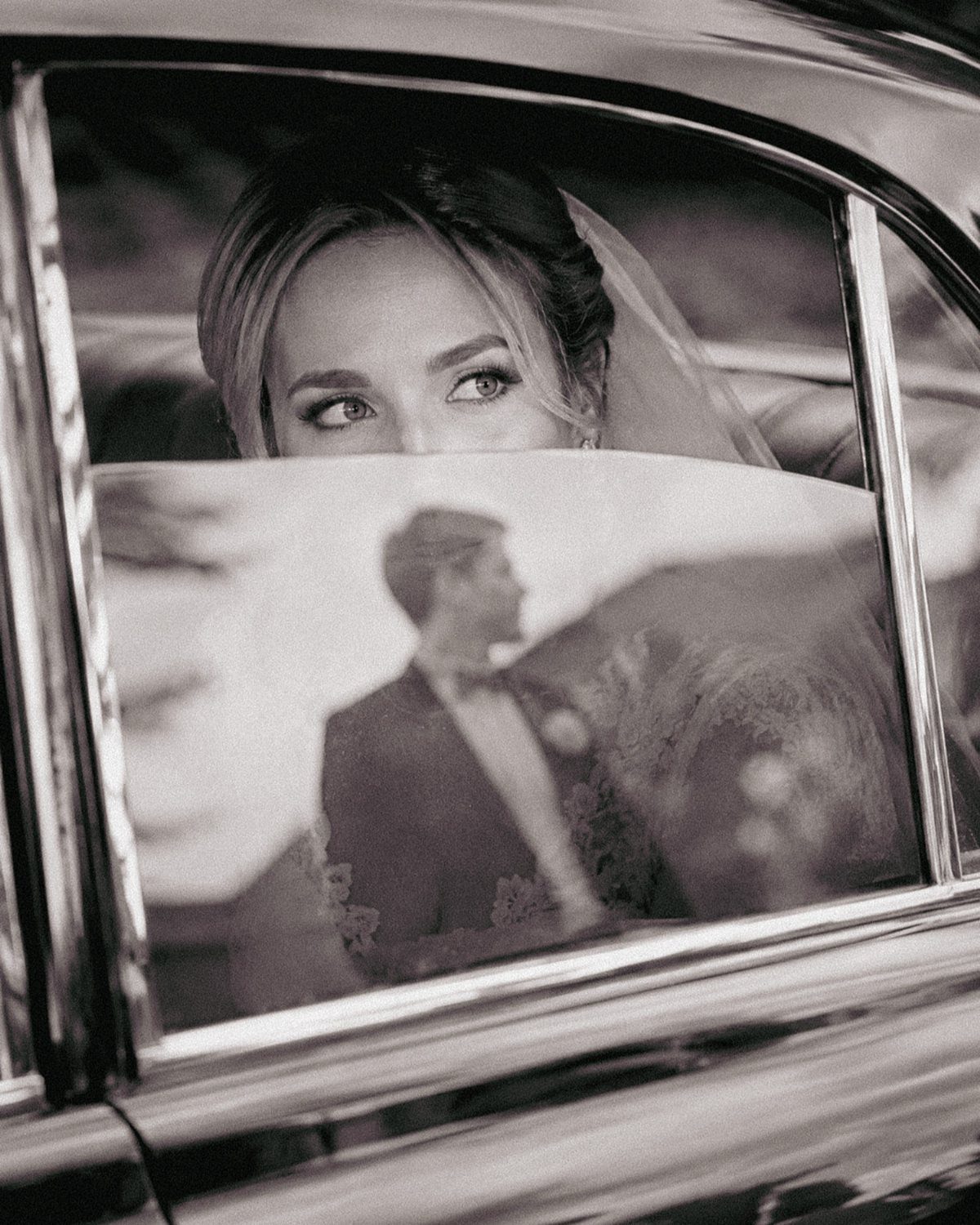 Bride looking through a car window with a veil and groom's reflection.