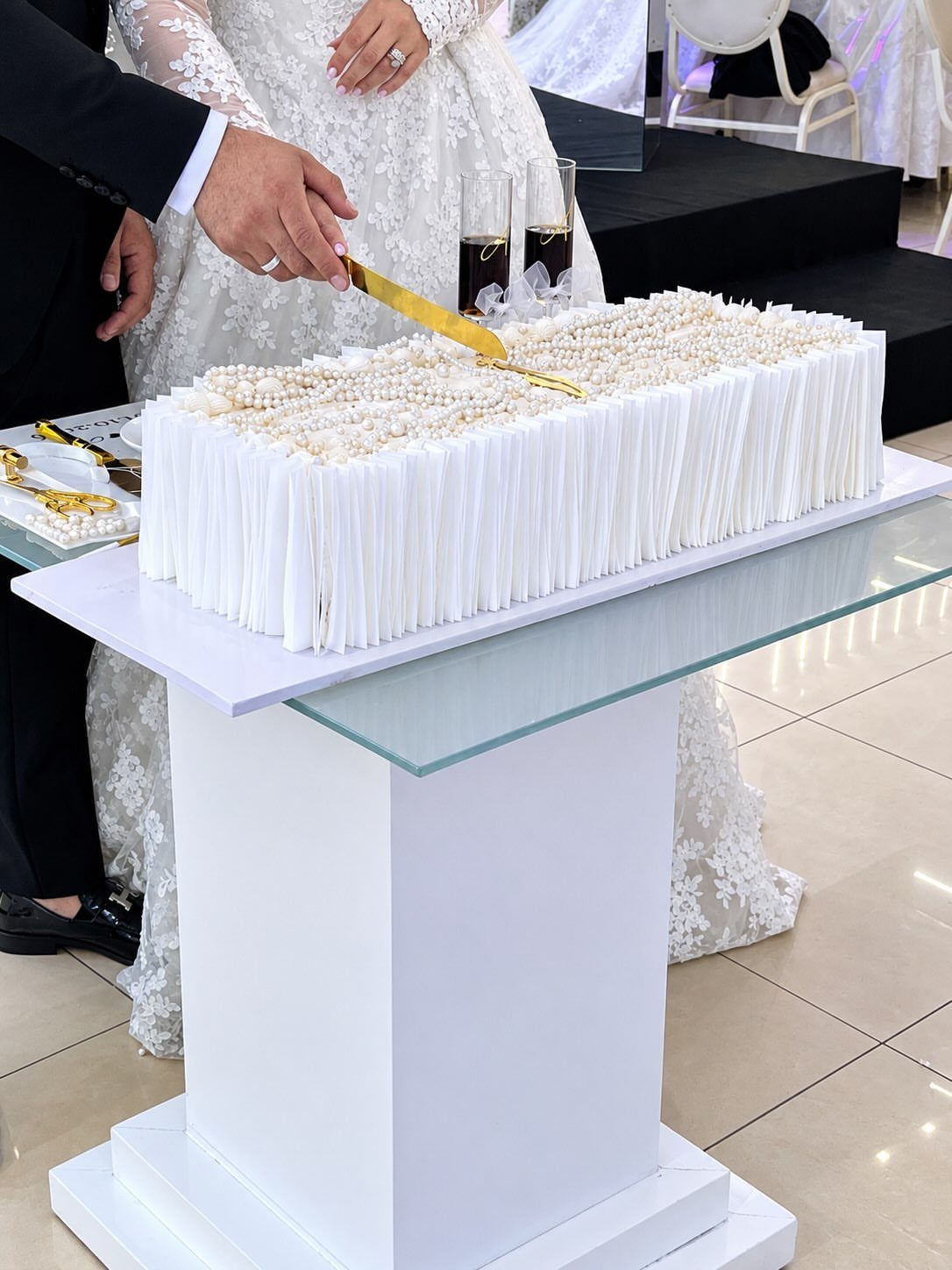 Couple in wedding attire cutting a white pearl-decorated cake on a white pedestal.