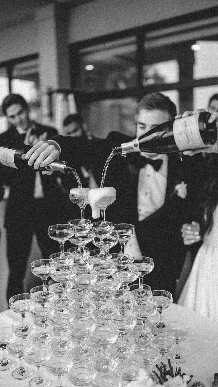 Two men in tuxedos pouring champagne into a tiered tower of coupe glasses.