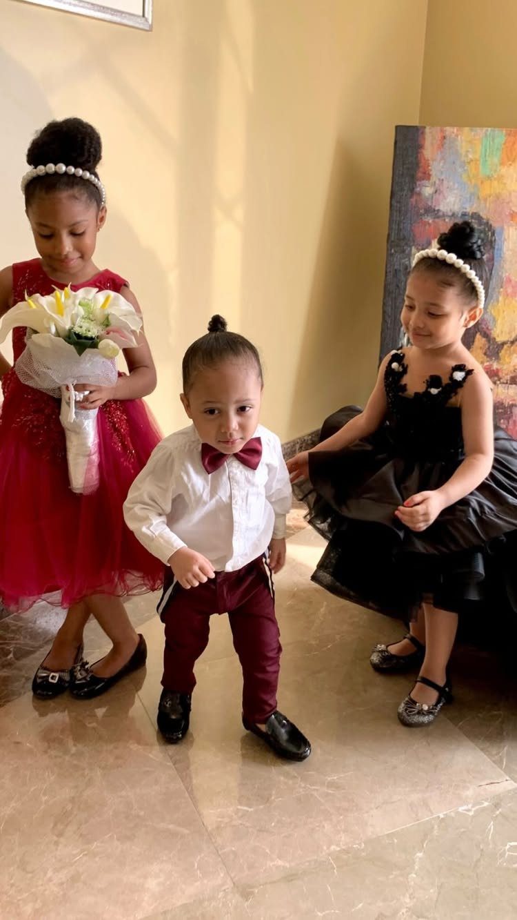 Three children in formal wedding attire posing indoors with a small bouquet.