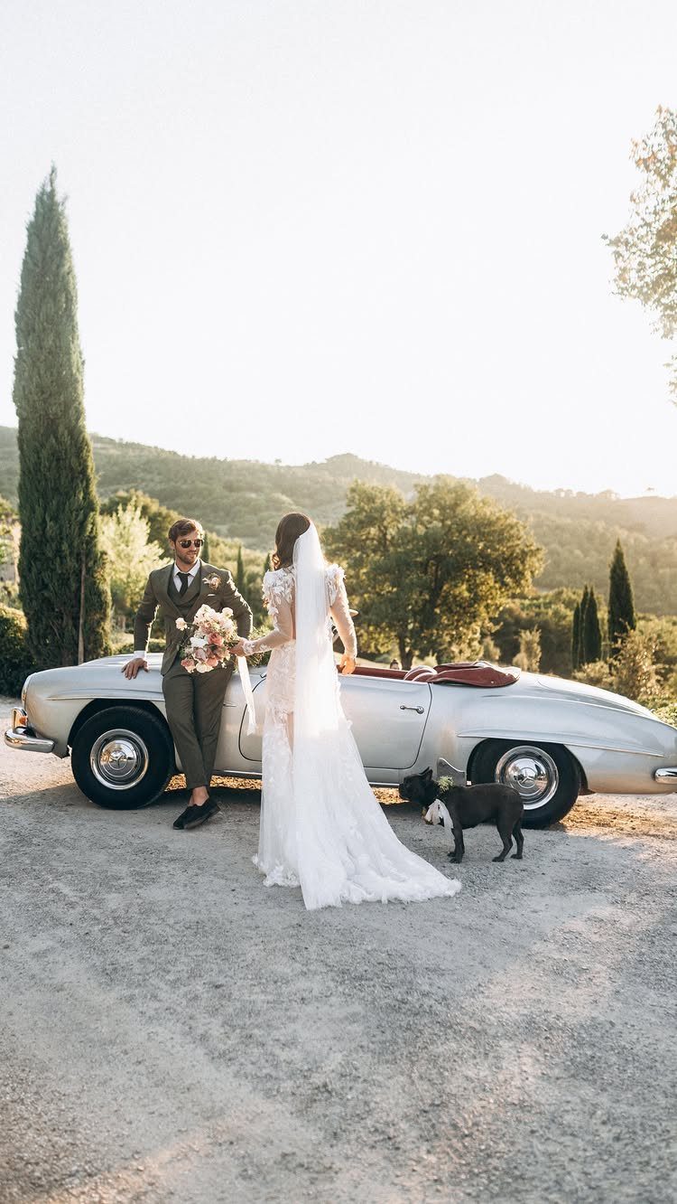 Bride and groom standing beside a silver vintage convertible car with a small dog.