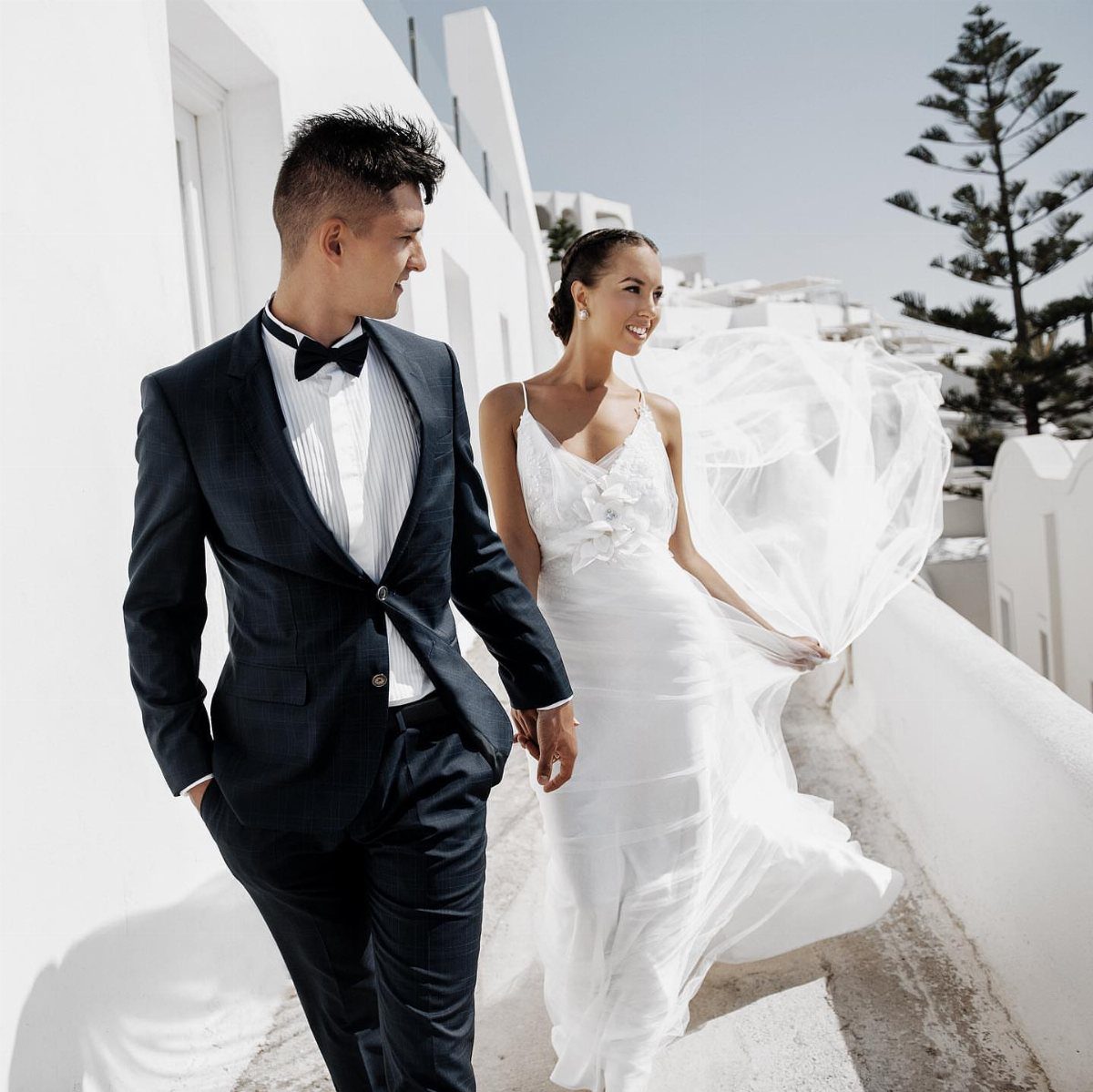 Bride and groom walking hand-in-hand through a white village with flowing veil.