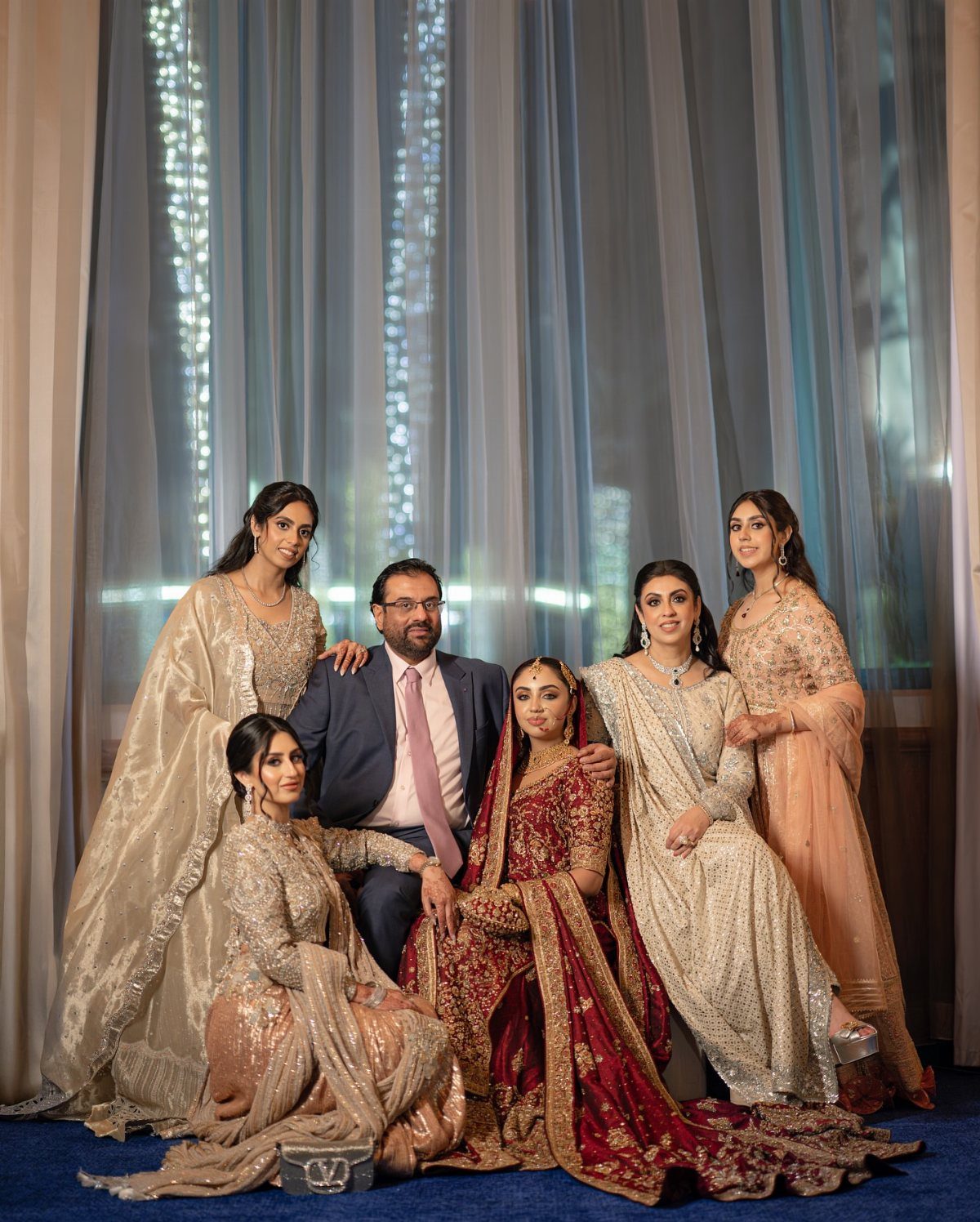 Family group in formal South Asian wedding attire posing against sheer blue curtains.