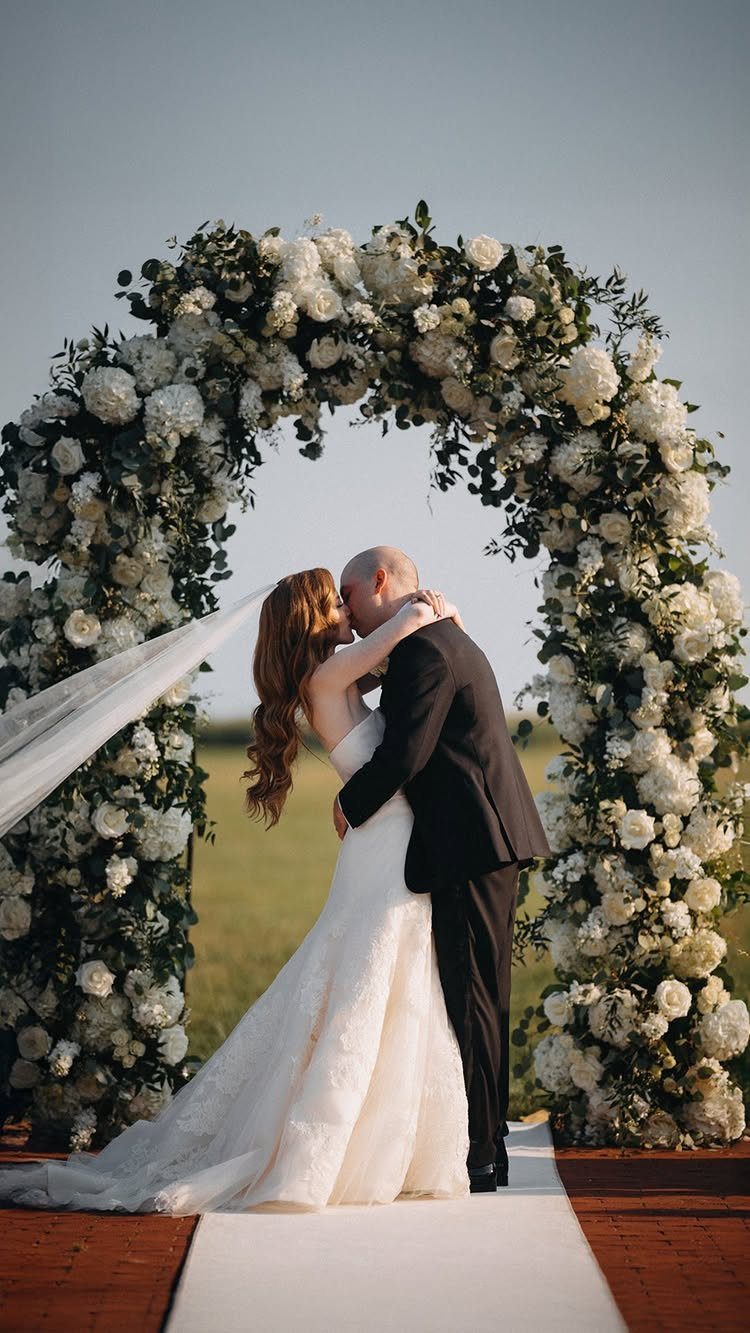 Bride and groom kissing under a white floral arch on a wooden deck.