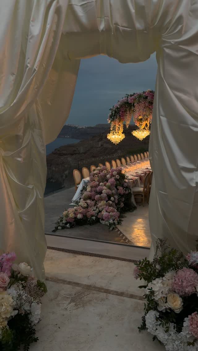 Outdoor wedding reception table with pink flowers and chandeliers overlooking the sea at dusk.