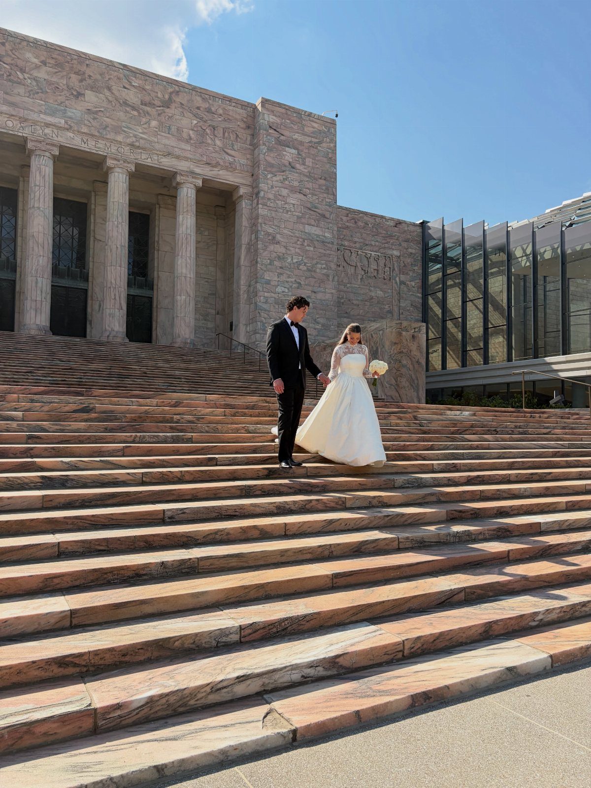Bride and groom walking hand-in-hand down wide marble steps of a stone building.