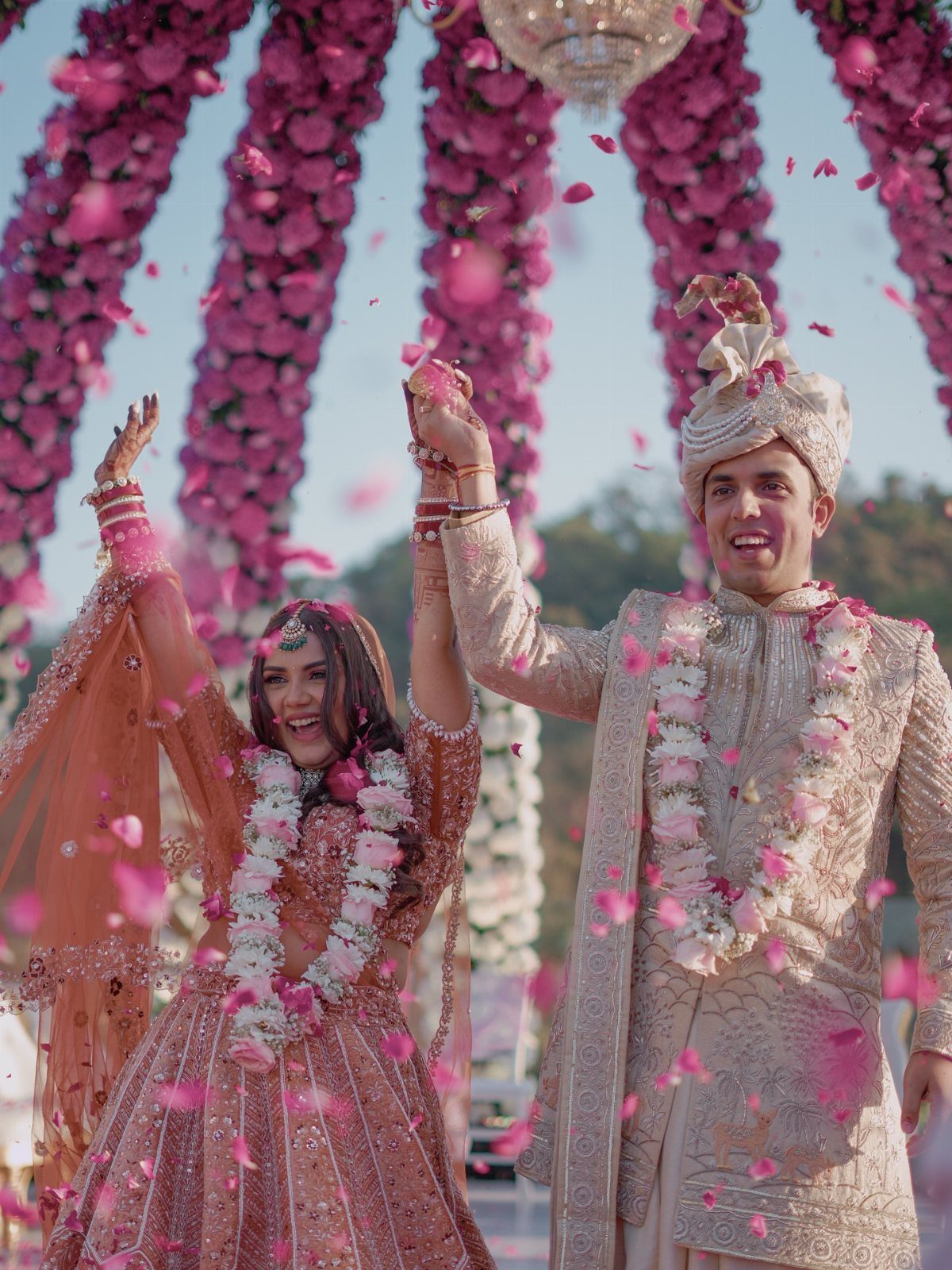Newlyweds in traditional Indian attire holding hands high while pink petals fall around them.