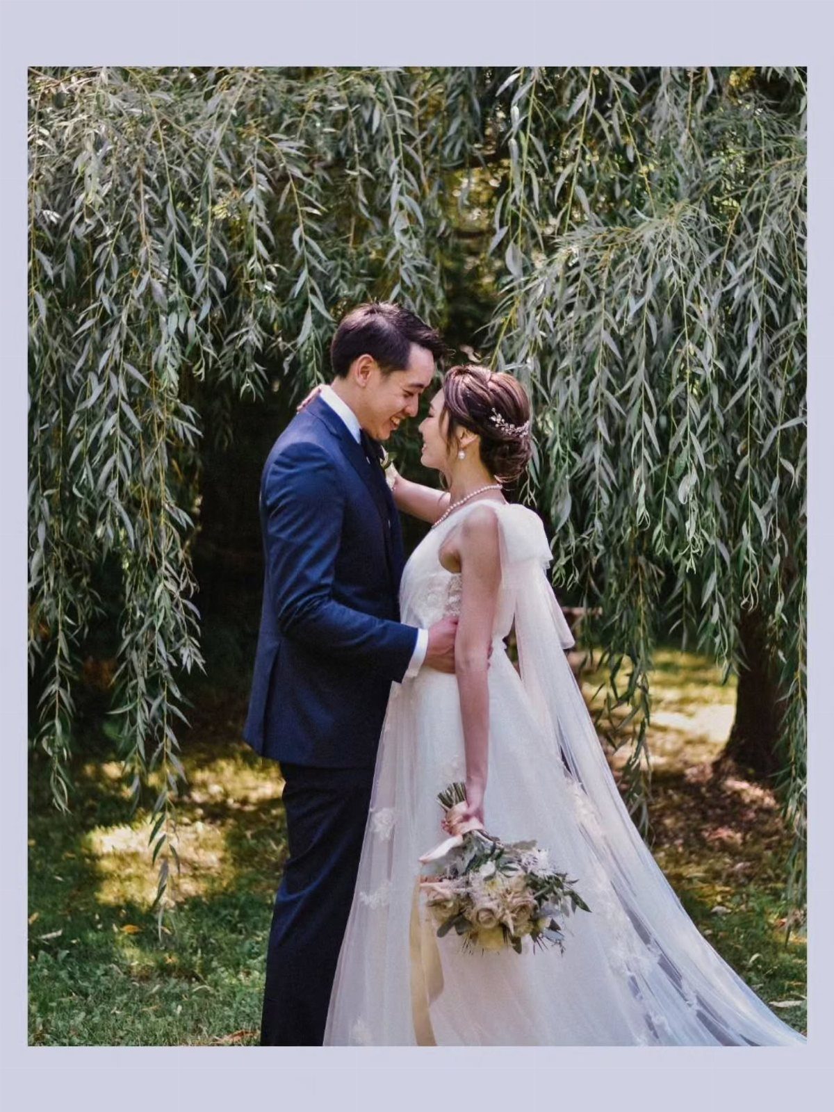 Bride and groom smiling face to face under a weeping willow tree.