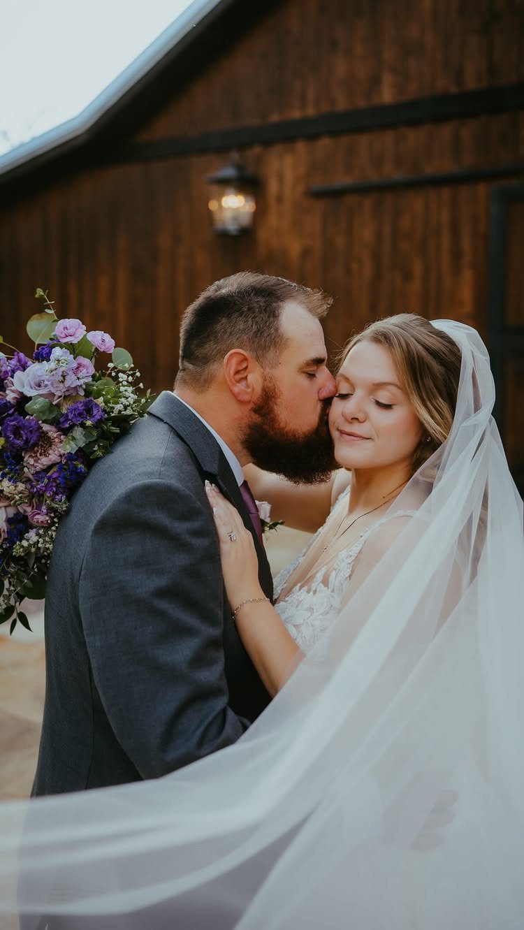 Groom in grey suit kissing the cheek of a bride in a white veil.