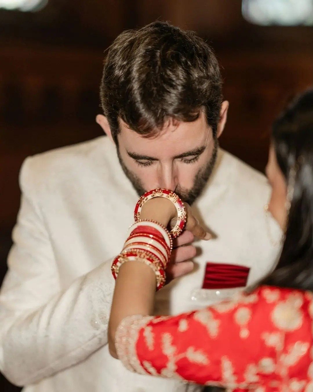Man in white suit kissing the hand of a woman in red attire.