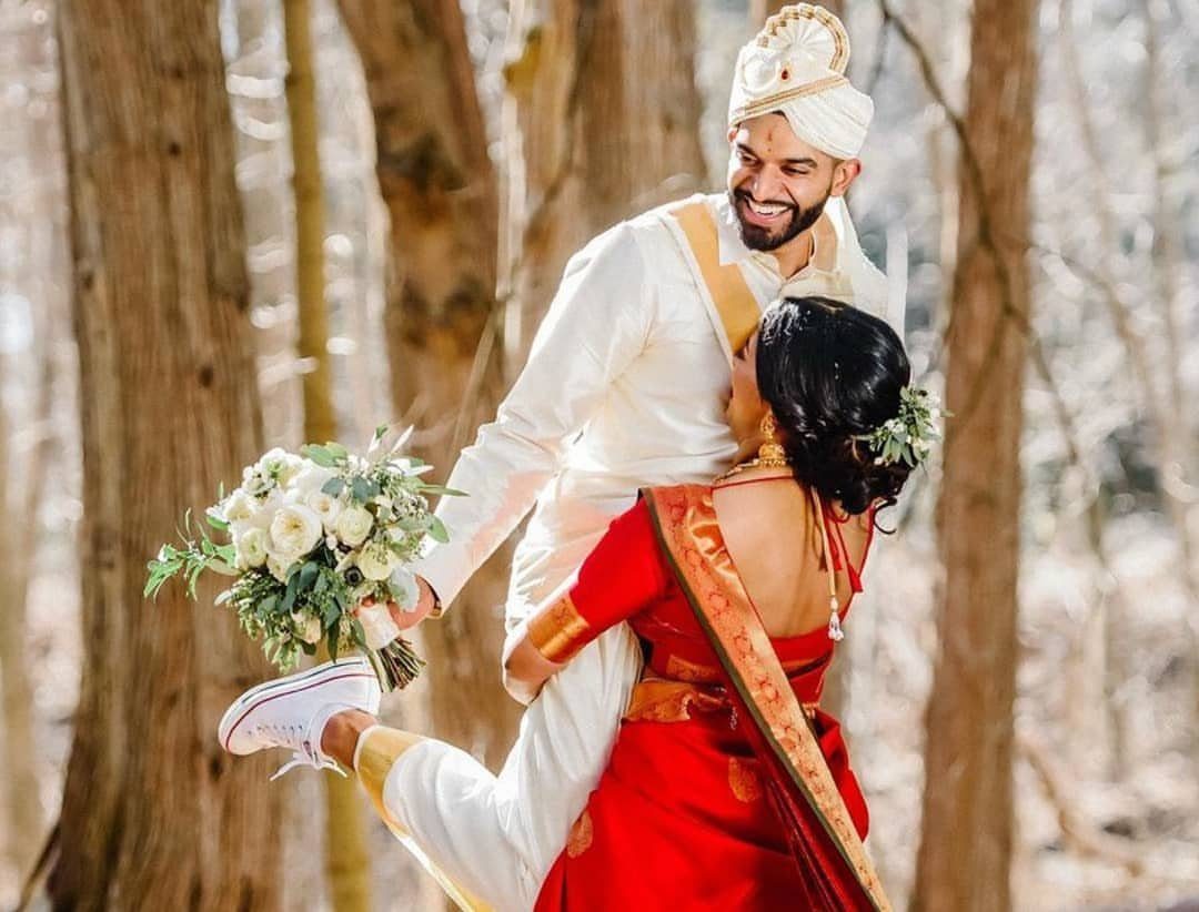 Groom lifting bride in red saree and white sneakers in a forest setting.