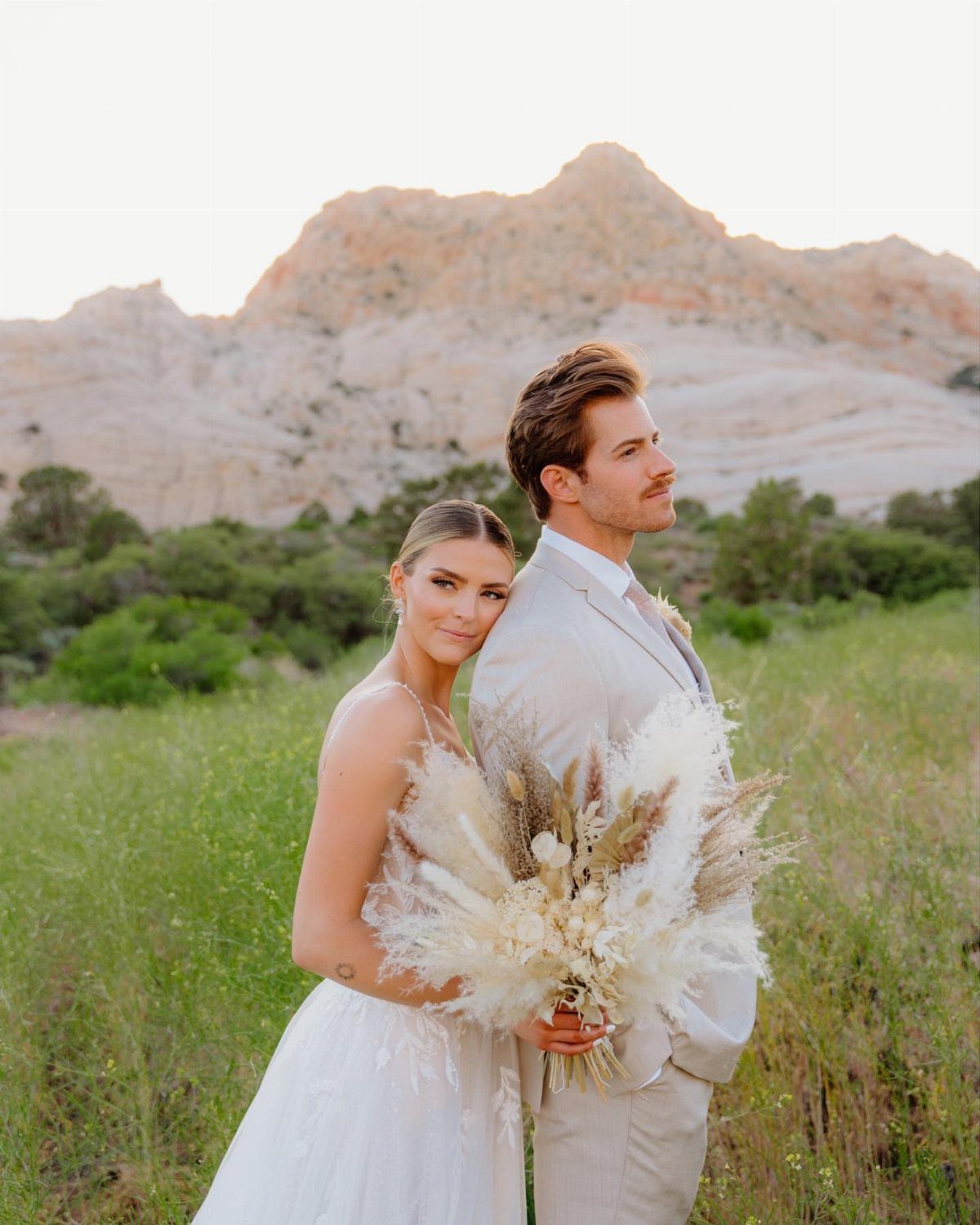 Bride and groom posing in a grassy field with rocky mountains behind them.