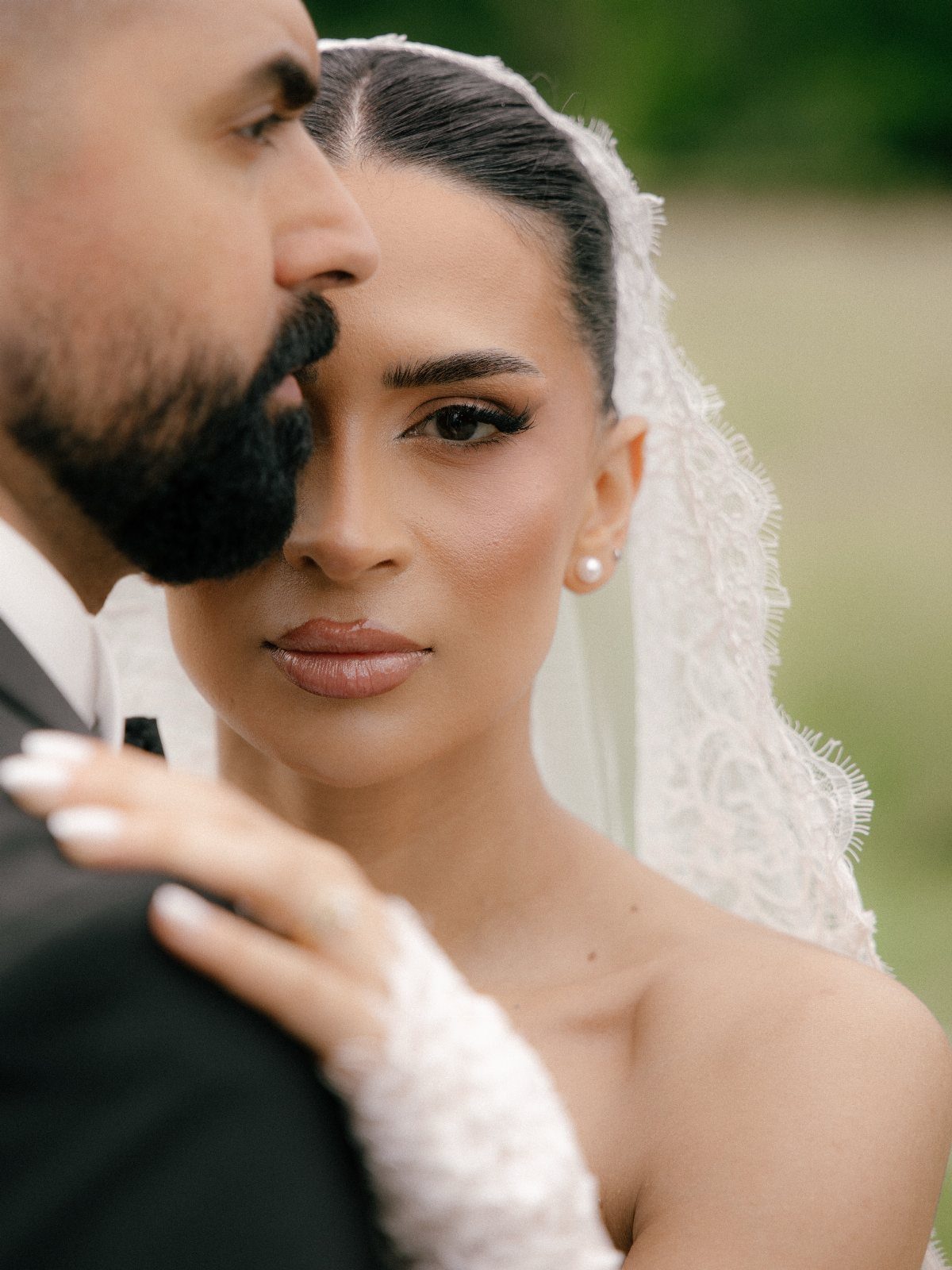 Bride in white lace veil looking at camera while leaning on groom