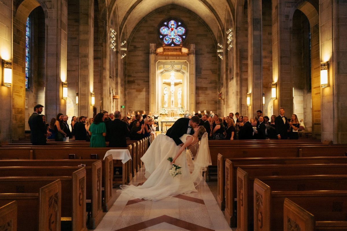 Groom dipping bride for a kiss in a cathedral aisle surrounded by guests.