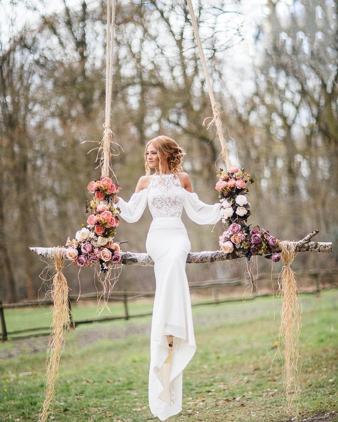 Bride in white lace dress standing on a flower-decorated wooden swing outdoors.
