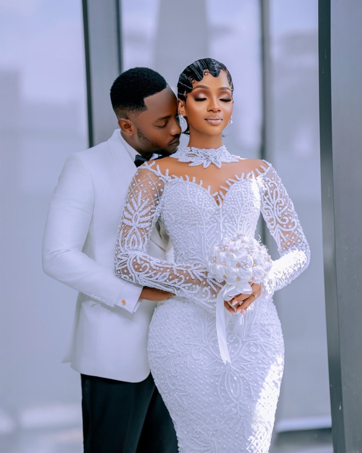 Groom in white tuxedo hugging bride from behind in beaded lace wedding gown