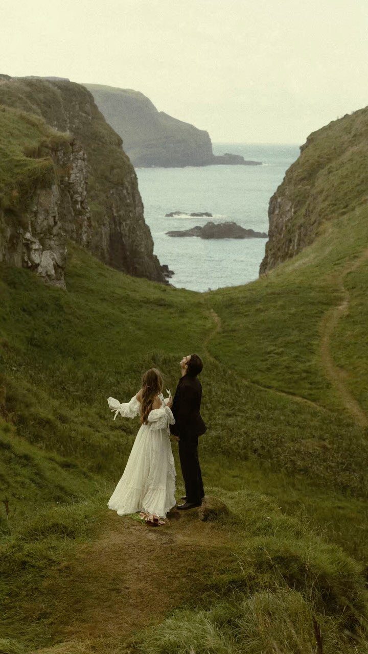 Wedding couple standing on a grassy cliffside path overlooking the ocean.
