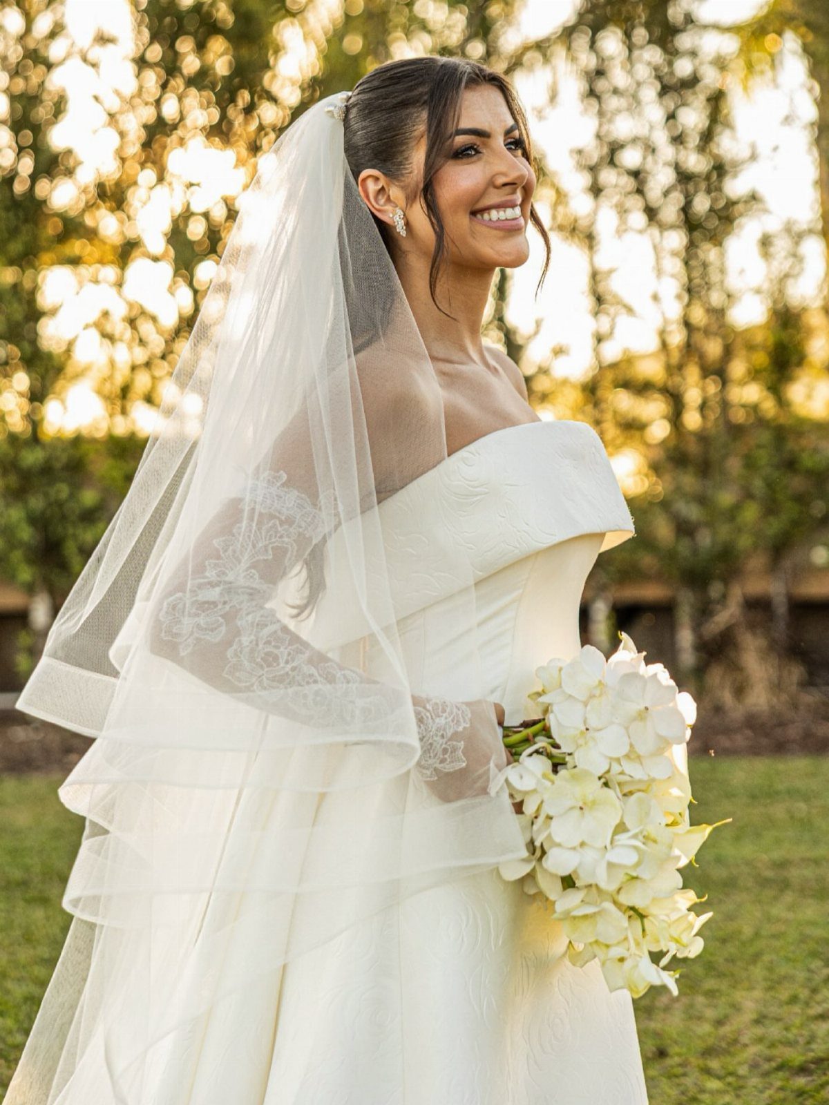 Smiling bride in white gown and veil holding white orchid bouquet outdoors.
