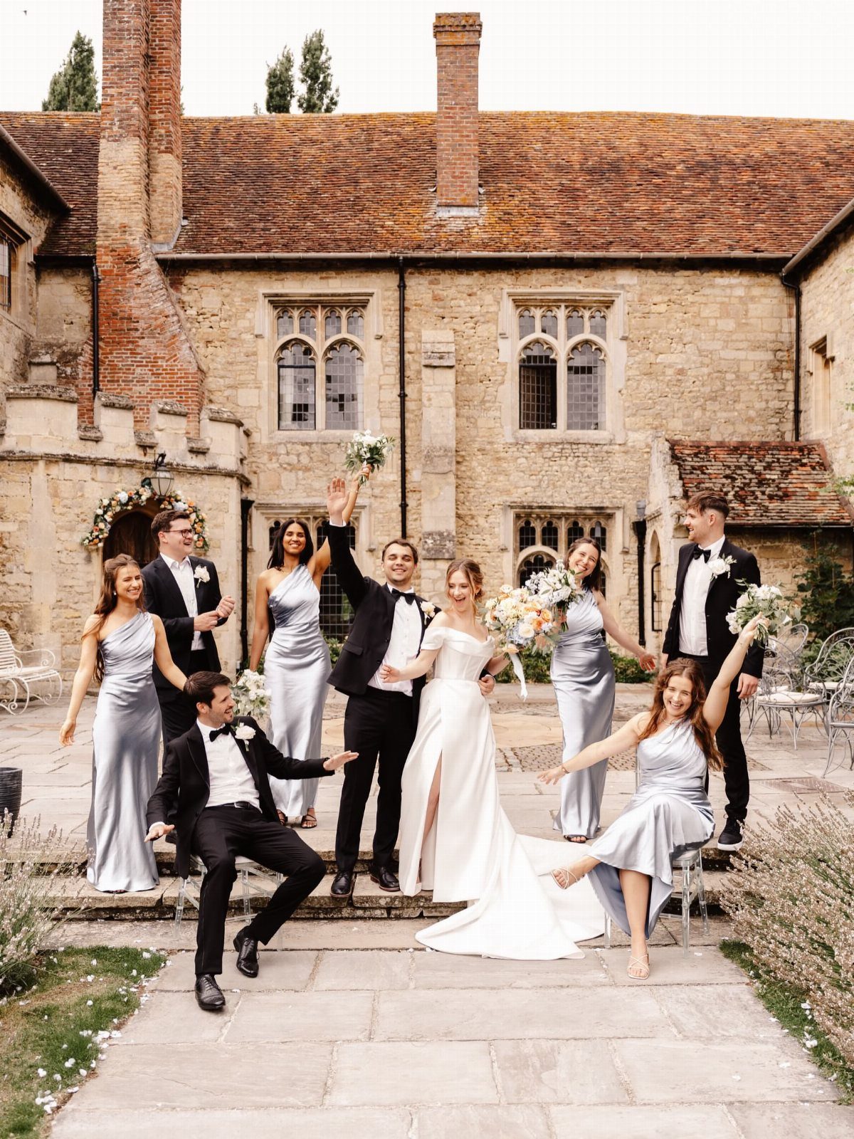 Bridal party posing playfully in front of a stone manor house with gardens.
