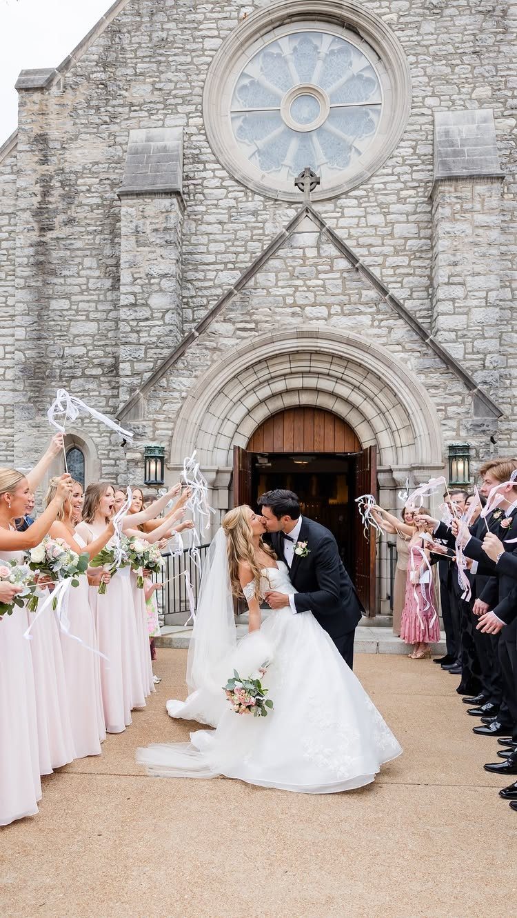 Bride and groom kissing outside a stone church while guests throw white ribbons.
