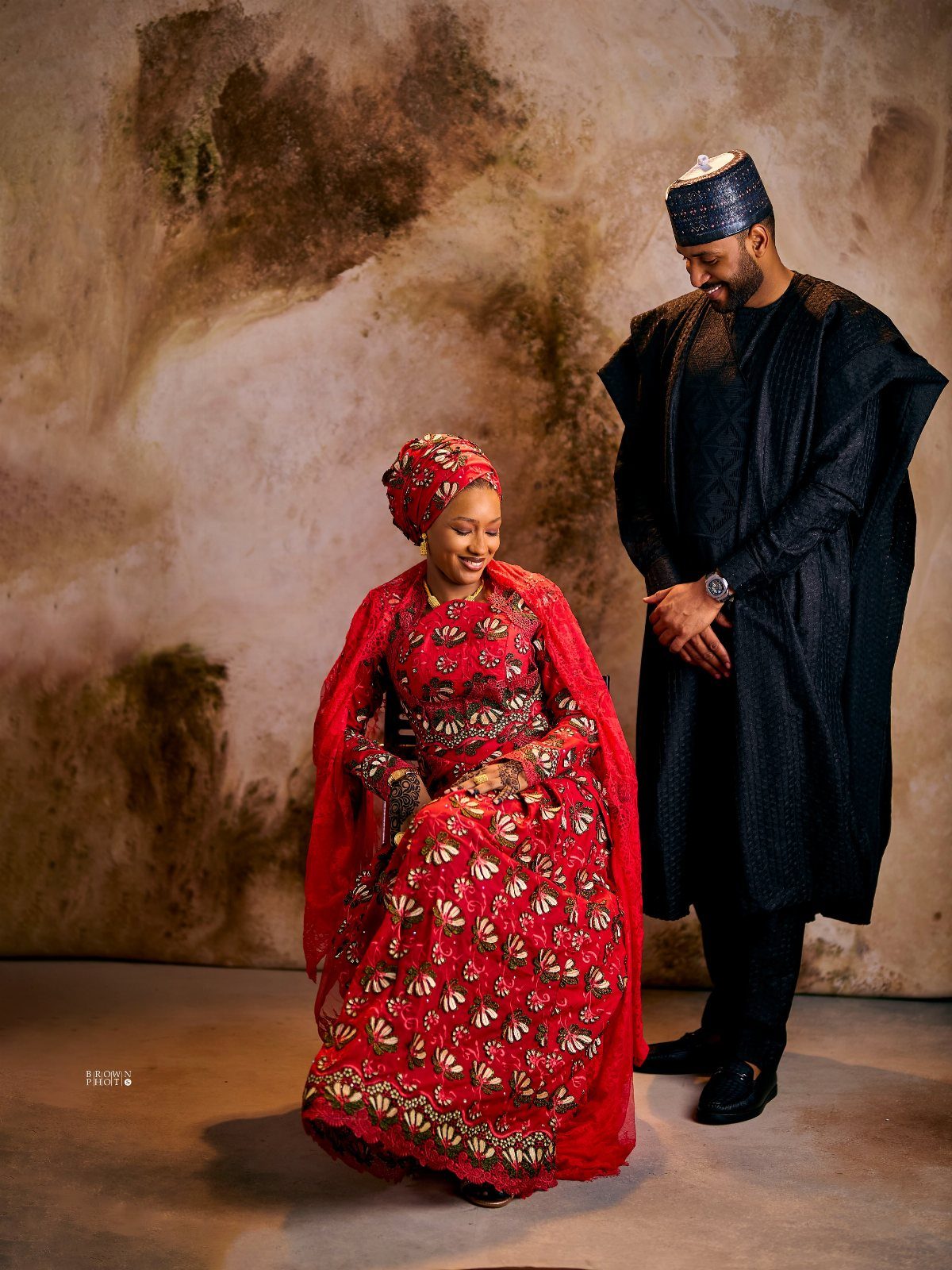 Man in black agbada standing beside a woman in red traditional bridal attire.