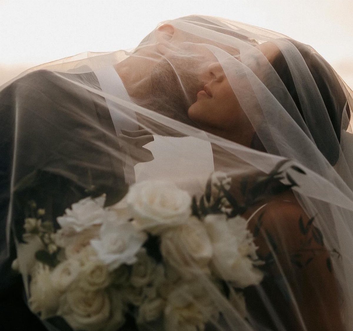 Bride and groom embracing under a sheer white veil with white roses.