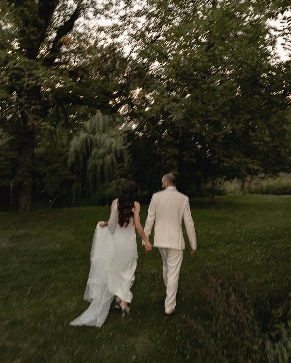 Bride and groom walking hand-in-hand away from camera through a green grassy field.