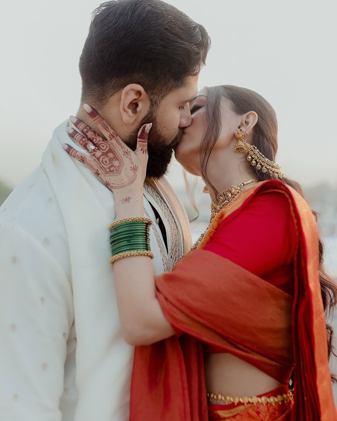 Couple in traditional Indian wedding attire kissing with henna on the woman's hand.