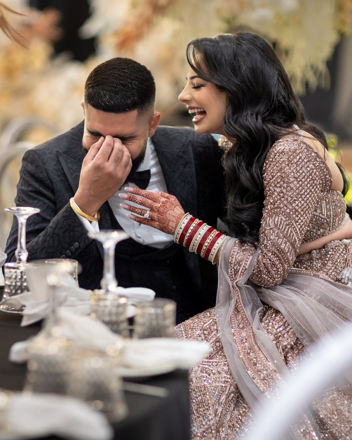 Couple in wedding attire laughing together while seated at a decorated reception table.