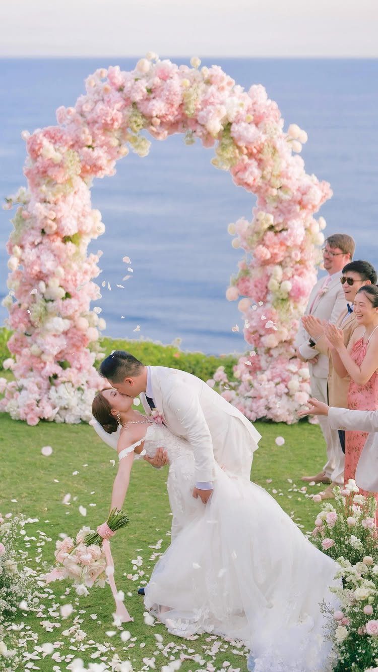 Groom dipping bride for a kiss under a pink floral arch by the ocean
