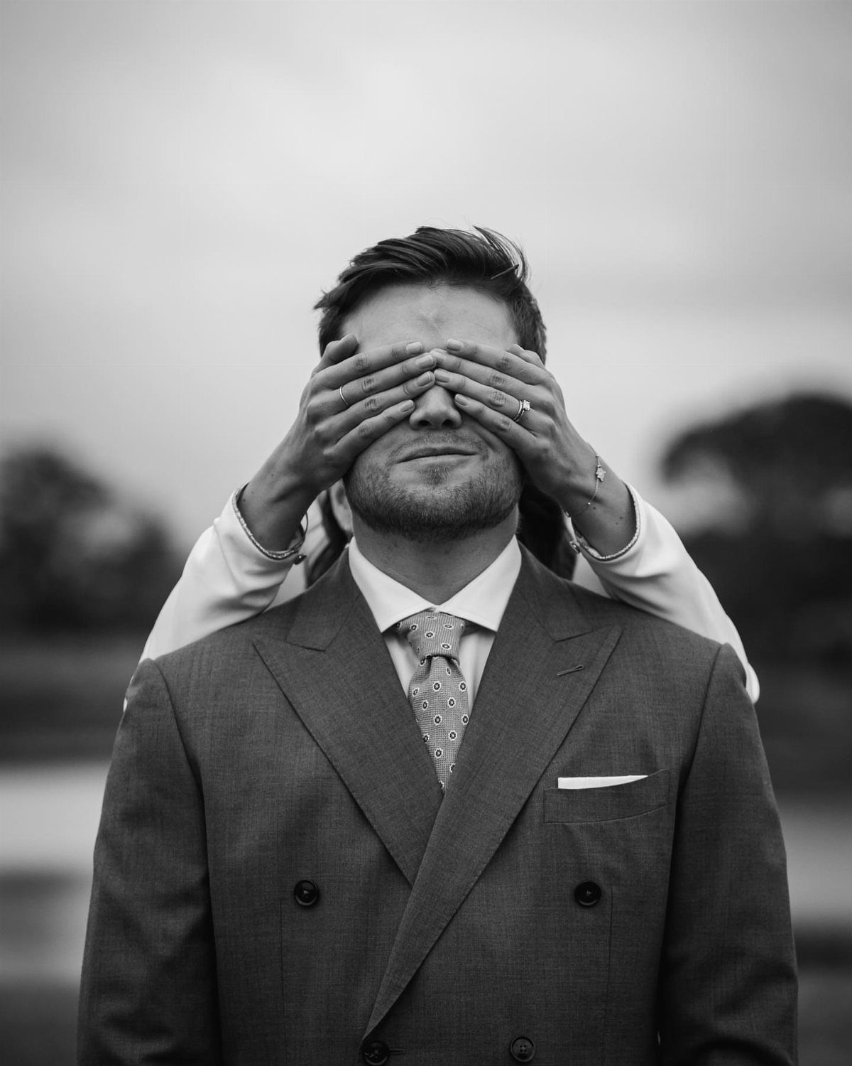 Groom in a suit with his eyes covered by a bride's hands.