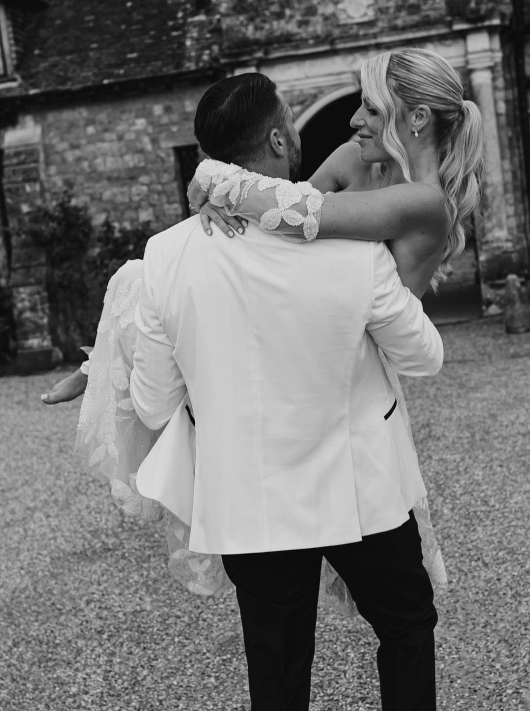 Groom in white tuxedo lifting bride in lace wedding dress outdoors
