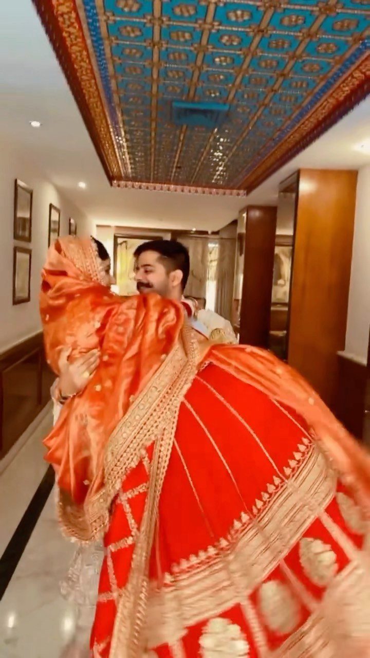 Groom lifting and swinging bride in red traditional Indian wedding attire in a hallway