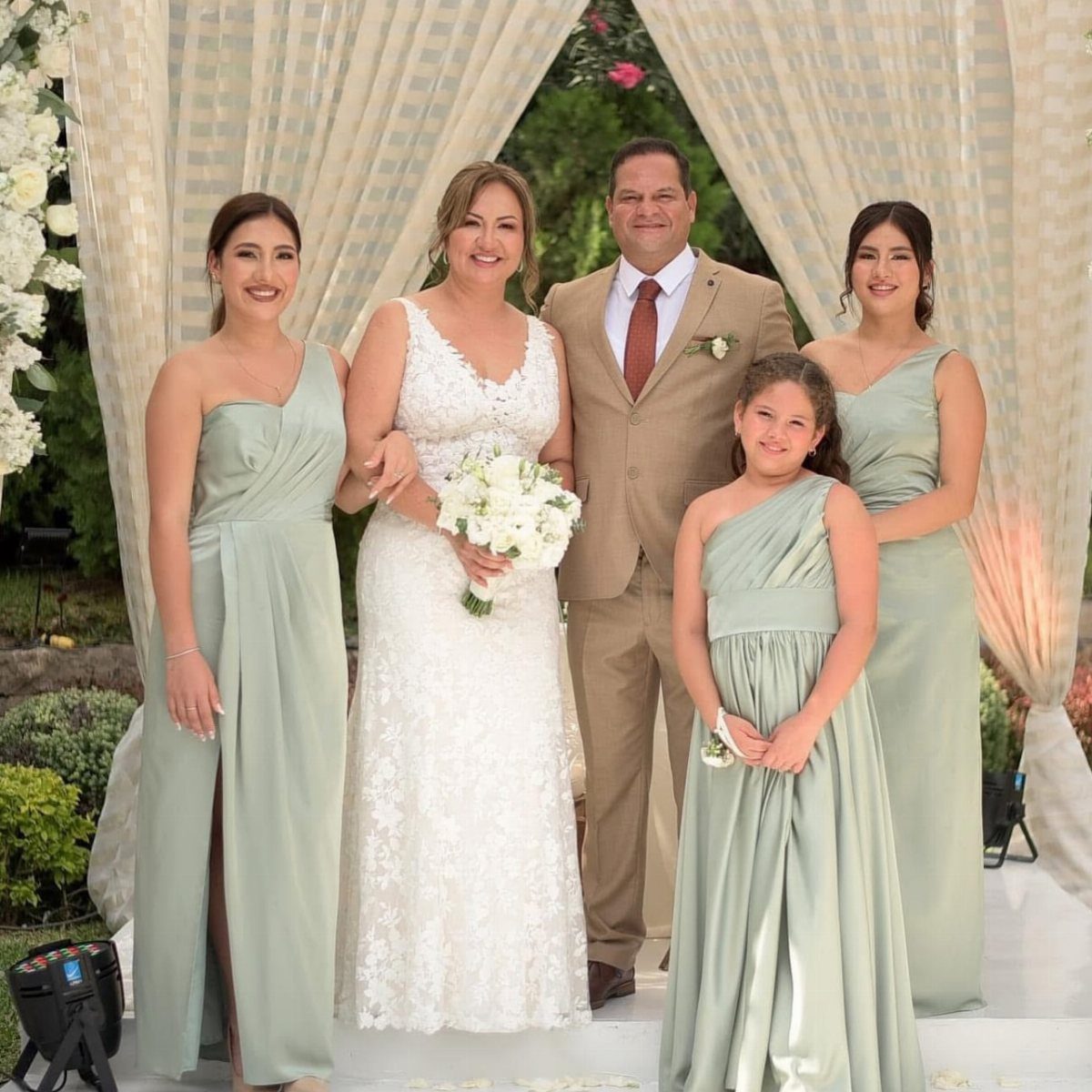 Bride and groom posing with bridesmaids and a flower girl under a draped arch