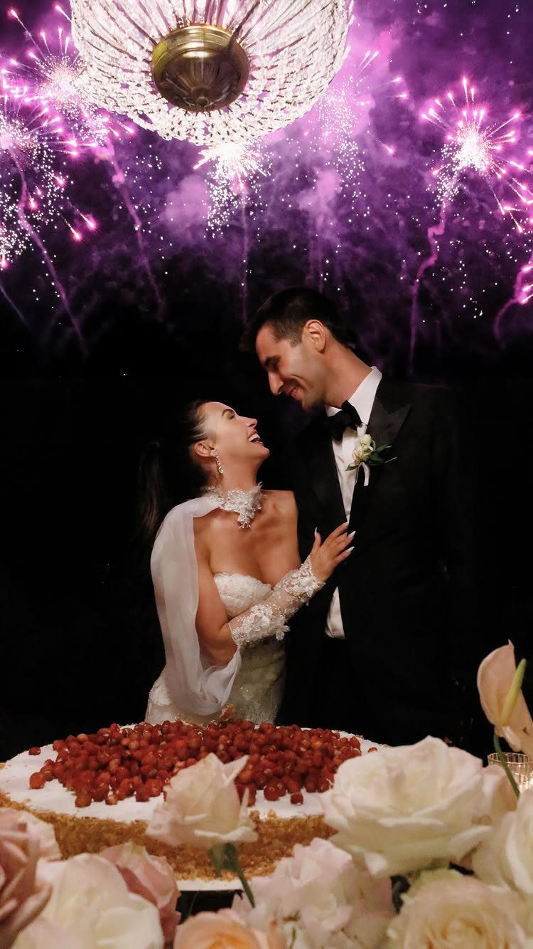 Bride and groom smiling at each other behind a cake with purple fireworks.