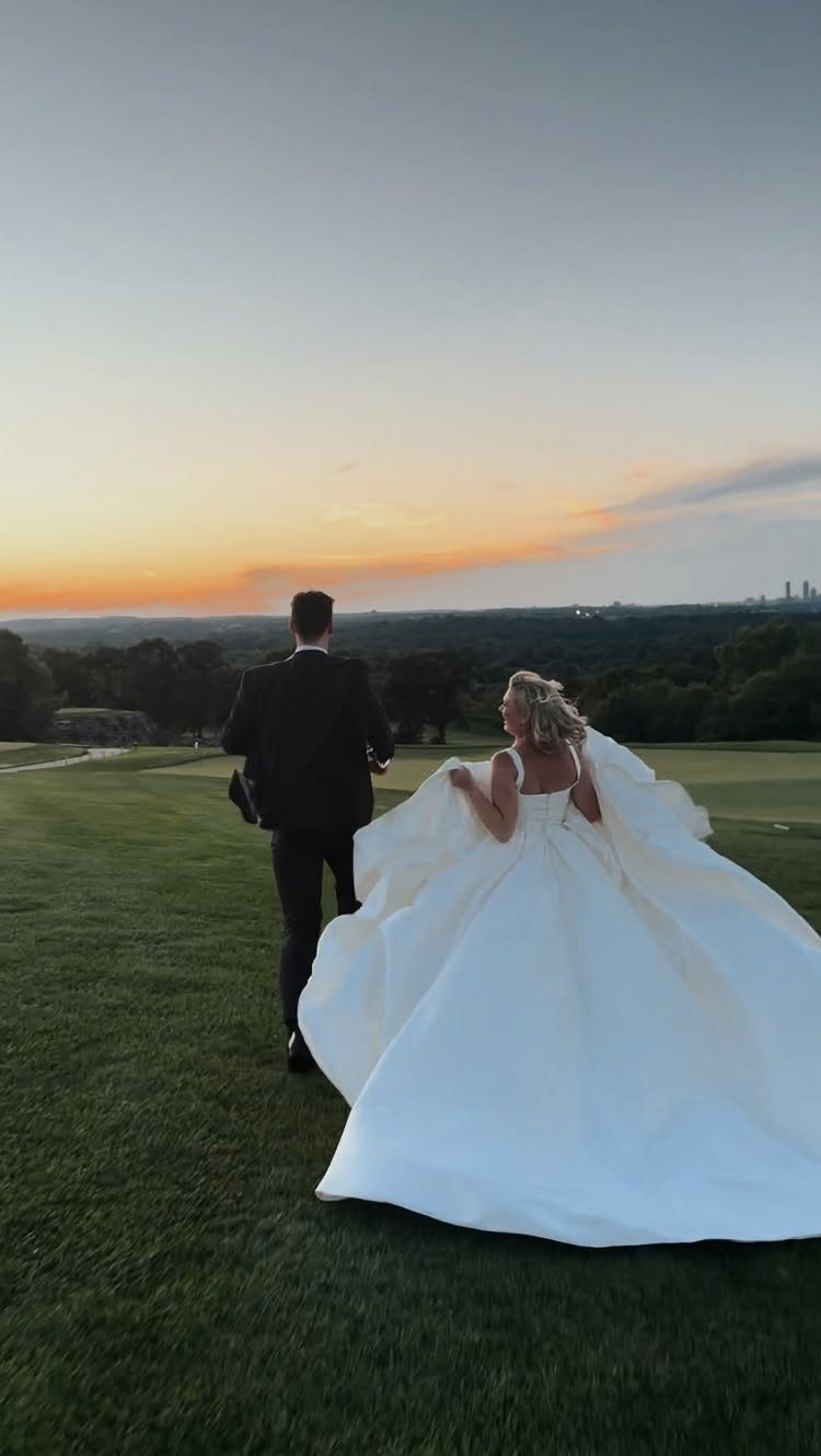 Bride and groom walking together across a green field during a golden sunset.