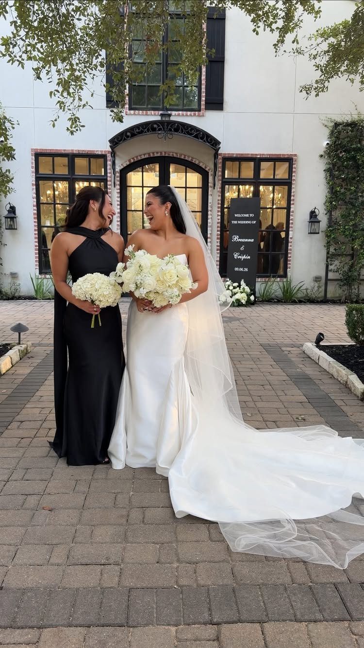 Two women in formal wedding attire standing side-by-side and smiling at each other.